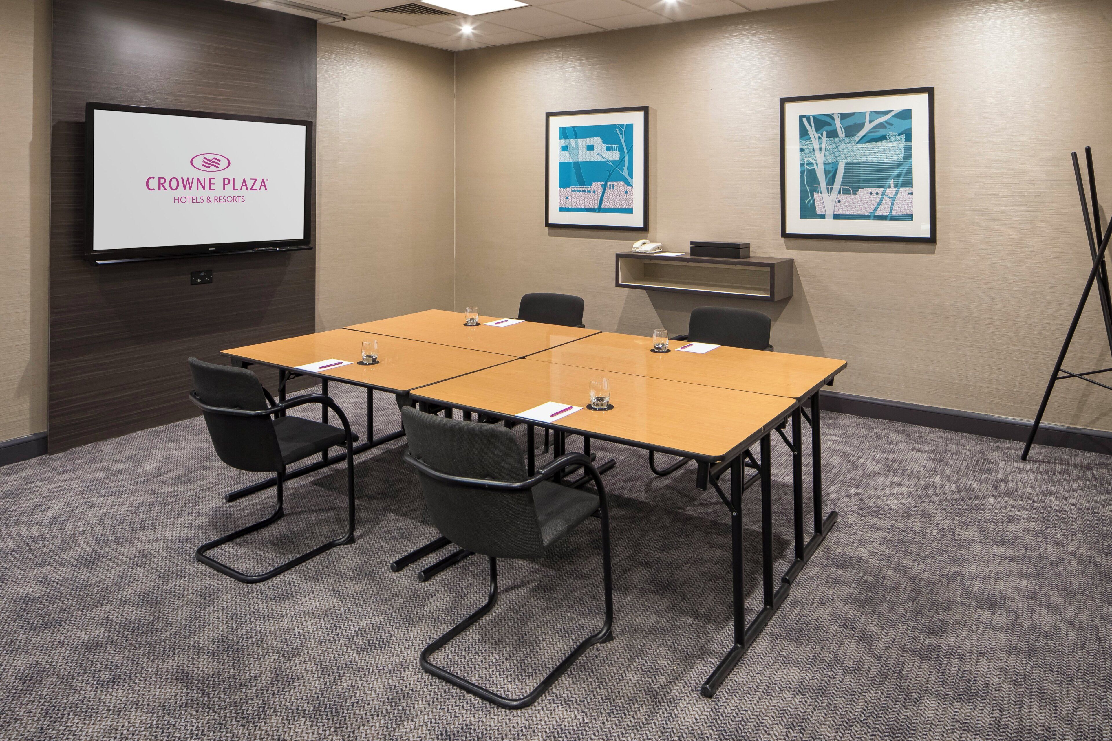 Een vergaderruimte met een groot scherm waarop het logo van 'CROWNE PLAZA HOTELS & RESORTS' staat, een houten tafel met vier stoelen, en twee ingelijste kunstwerken aan de muur.