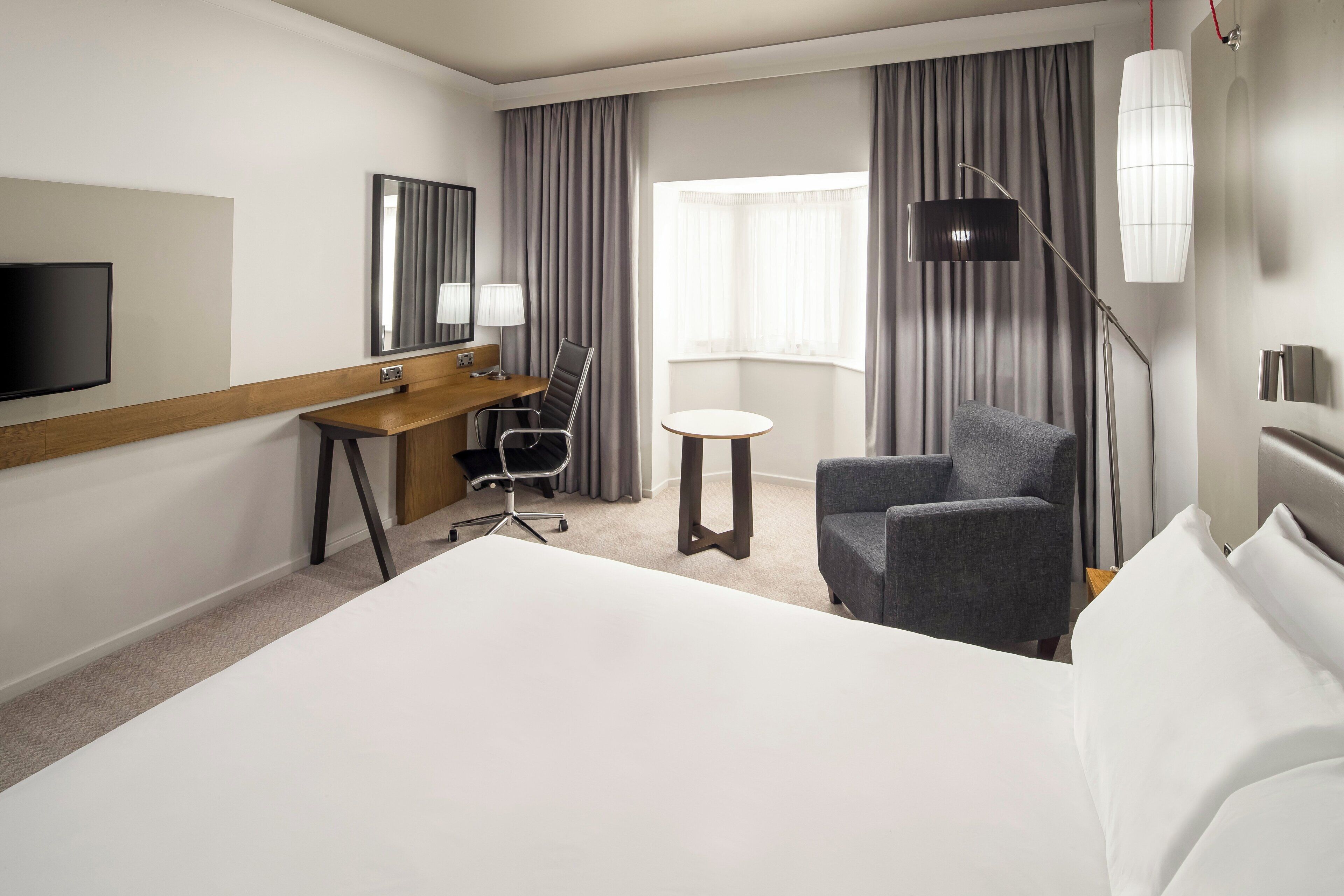 Una habitación de hotel con una cama grande, un escritorio con una silla, una mesita redonda pequeña, un televisor y una lámpara.