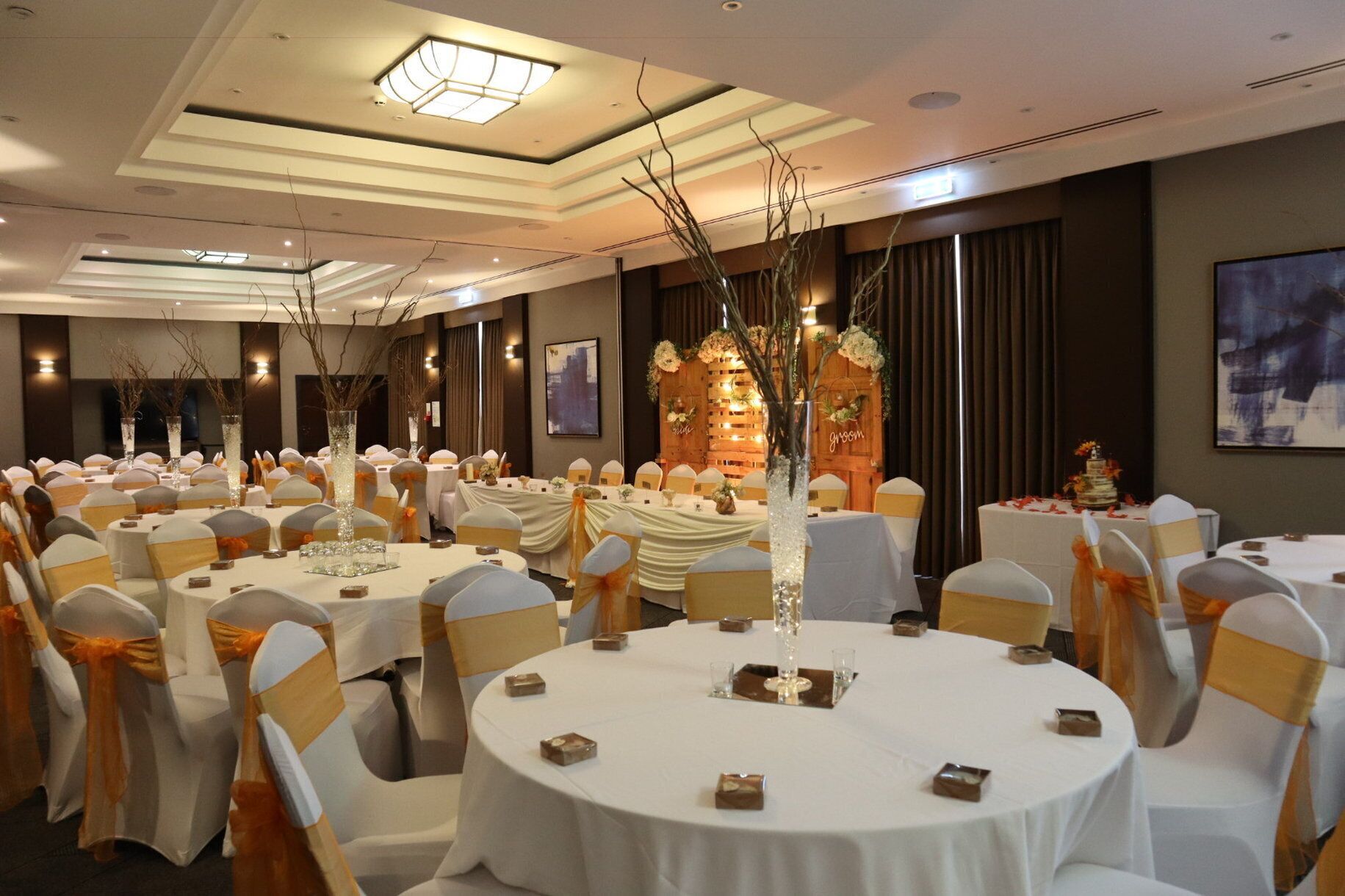 Un salón de banquetes con mesas redondas, sillas adornadas con cintas anaranjadas y un centro de mesa decorativo.