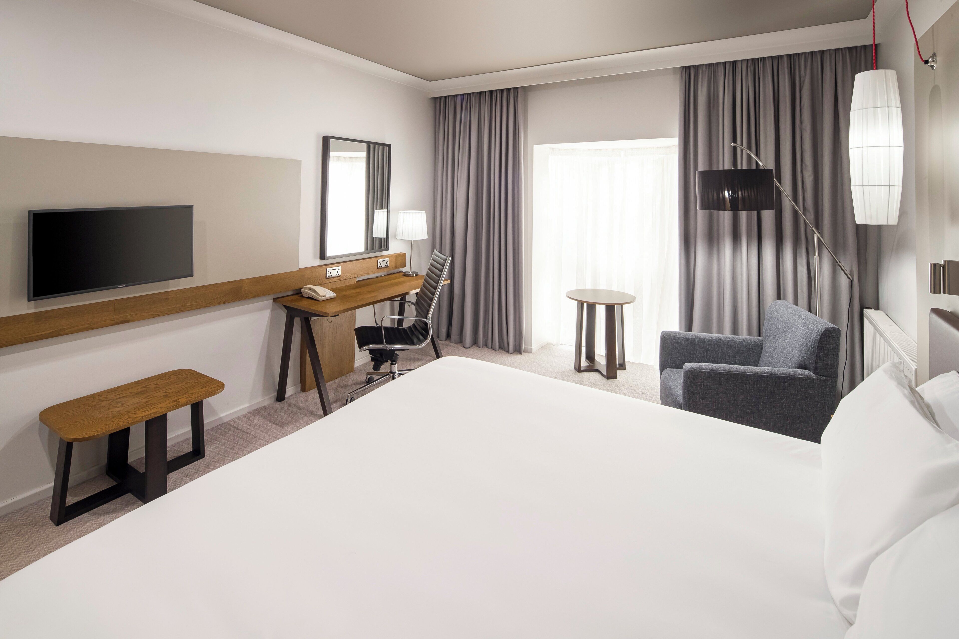 A hotel room with a large bed, a desk with a telephone, a chair, a TV, and a lamp.