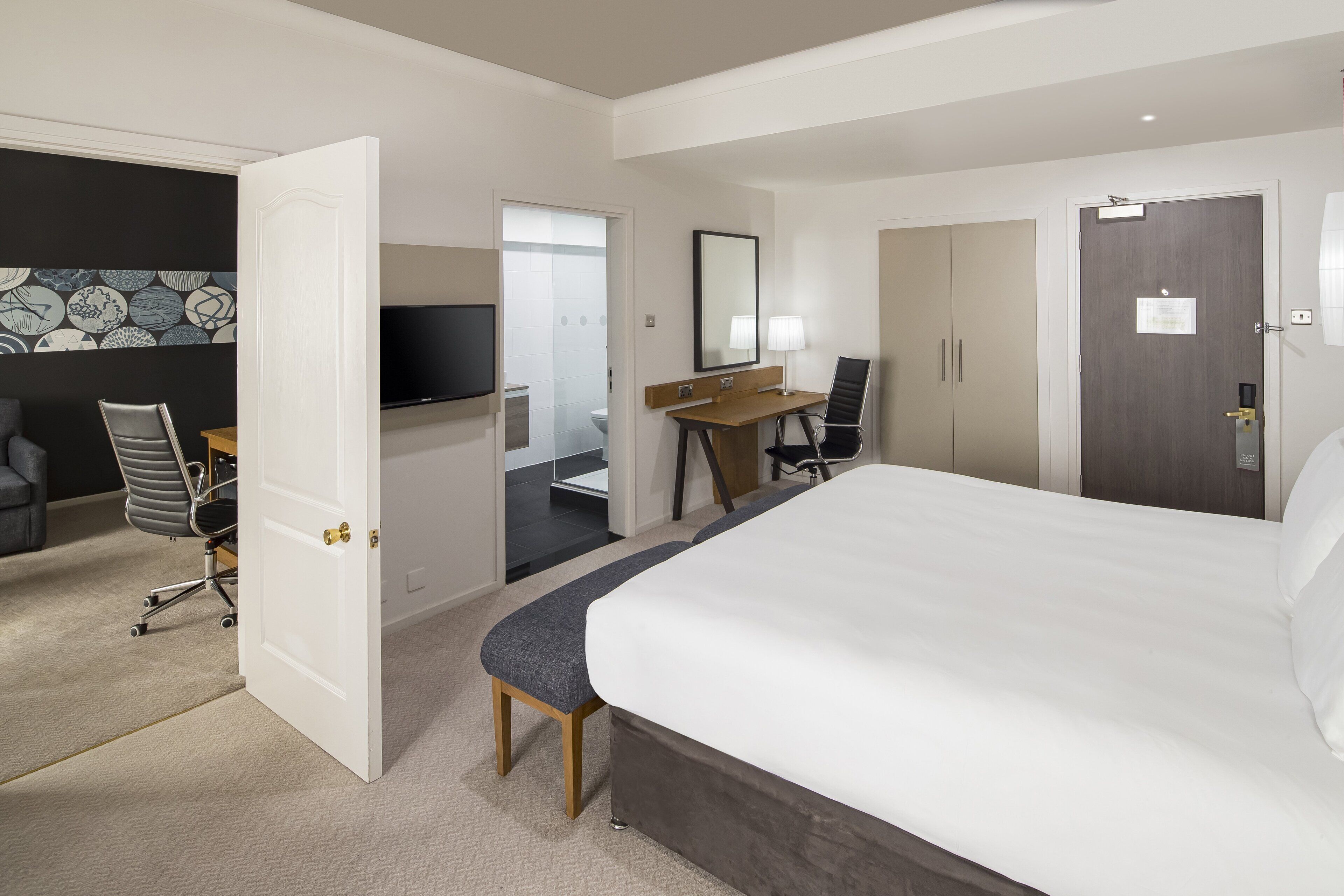 Une chambre d’hôtel avec un grand lit, un bureau avec une chaise, une télévision et une salle de bain visible à travers une porte ouverte.