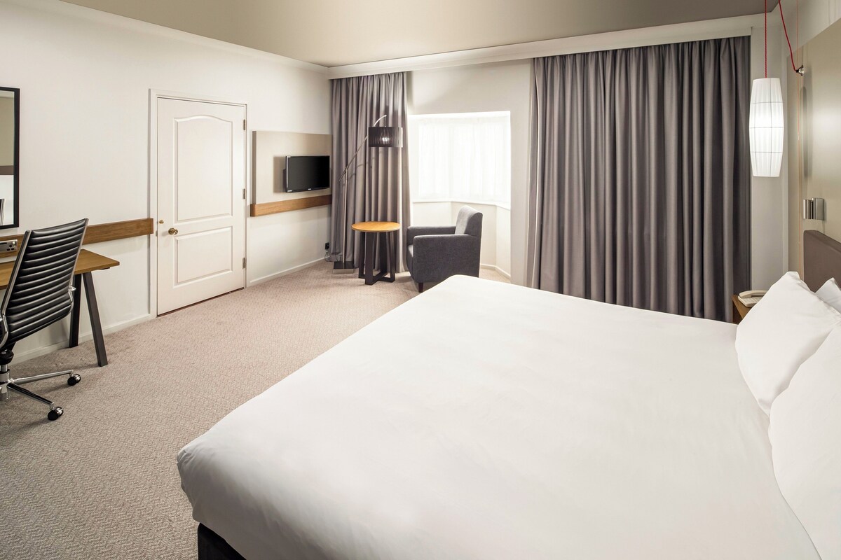 A hotel room with a large bed, a desk with a chair, a TV, and a window with curtains.