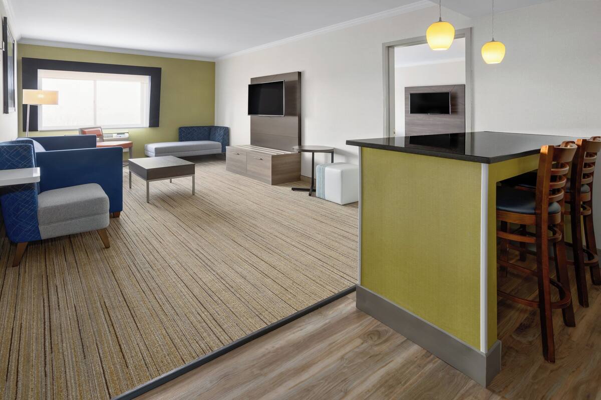A modern hotel room with a kitchenette, a seating area with a sofa and chairs, and a large window providing natural light.