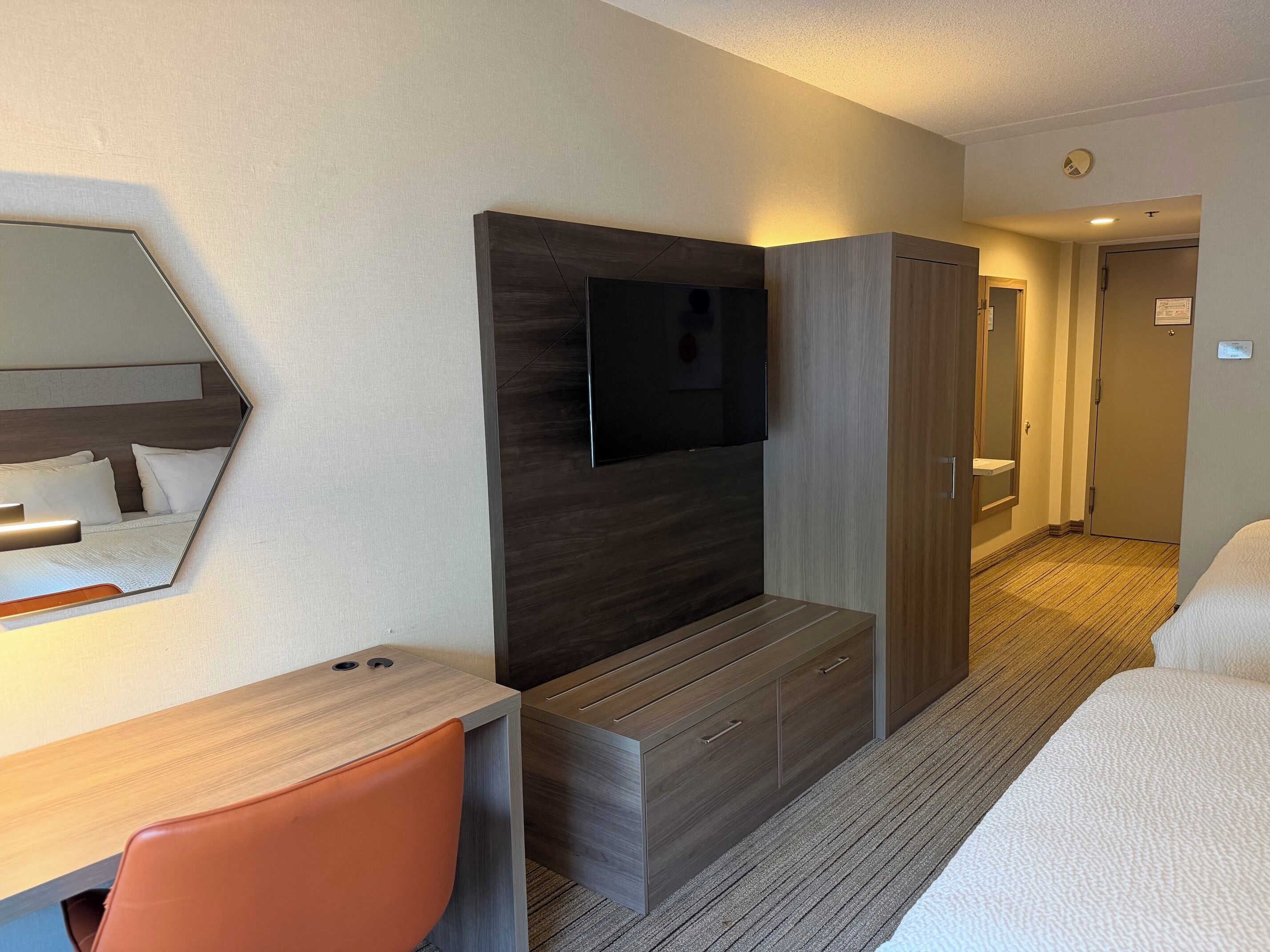 Quarto de hotel com cama, escrivaninha, cadeira, televisão e guarda-roupa.