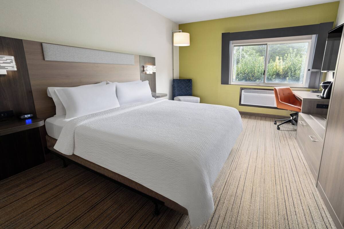 A hotel room with a large bed, a desk with a computer, a chair, and a window with a view of greenery.