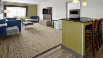 Una habitación de hotel moderna con kitchenette, un área de descanso con sofá y sillas, y un ventanal que proporciona luz natural.