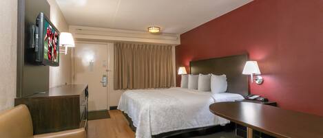 A hotel room with a bed, a desk, a chair, a TV, and a lamp.