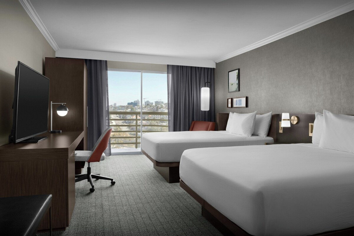 Habitación de hotel con dos camas, un escritorio con televisor, una silla y vistas a la ciudad a través de amplios ventanales.