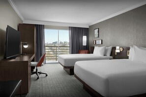 A hotel room with two beds, a desk with a TV, a chair, and a view of the city through large windows.