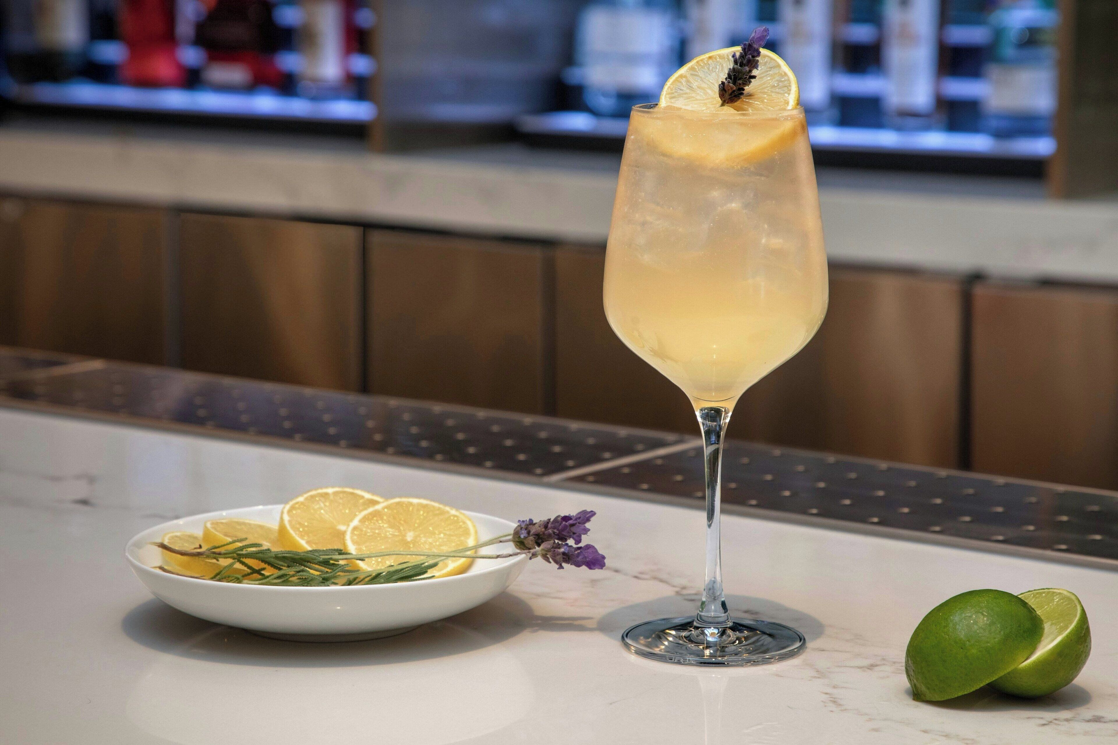 A cocktail glass with a lemon slice garnish, a plate with lemon slices and rosemary, and a lime on a marble countertop.