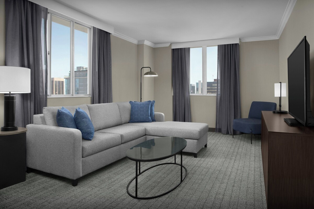Una habitación de hotel moderna con un sofá seccional gris, una mesa de centro de vidrio, un televisor de pantalla plana y un escritorio con una lámpara.
