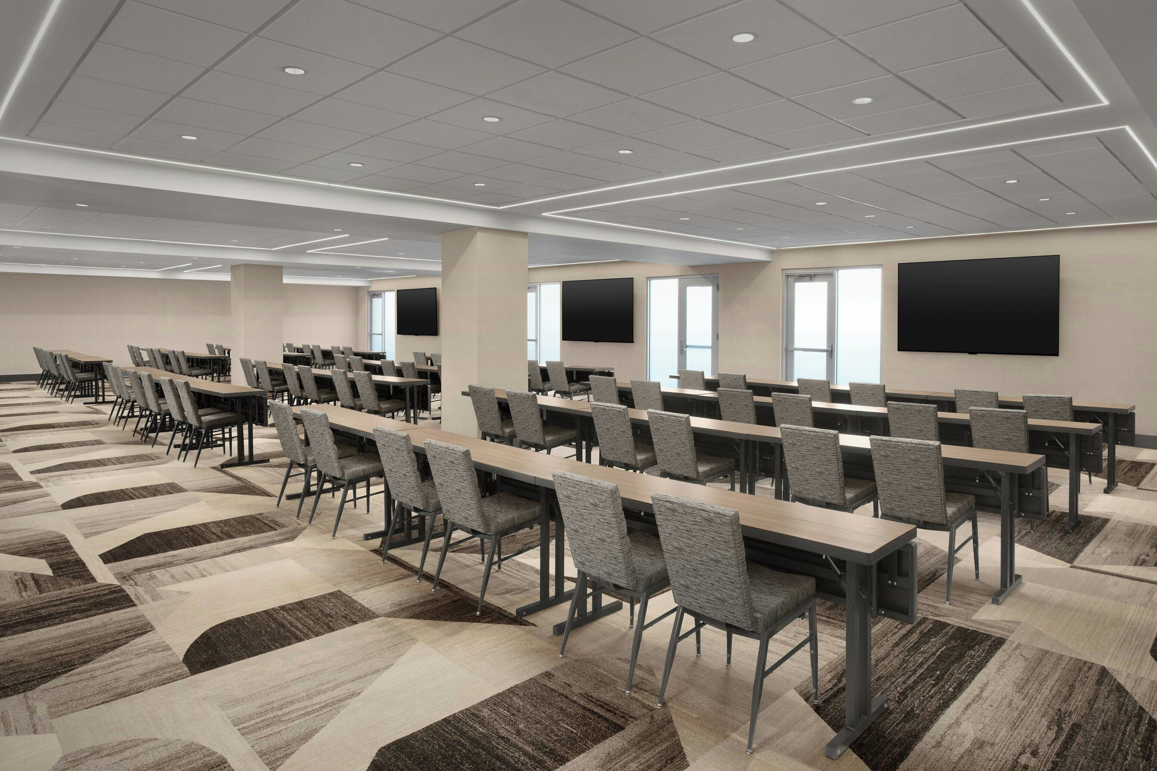 A conference room with rows of tables and chairs, large windows, and a flat-screen TV on the wall.