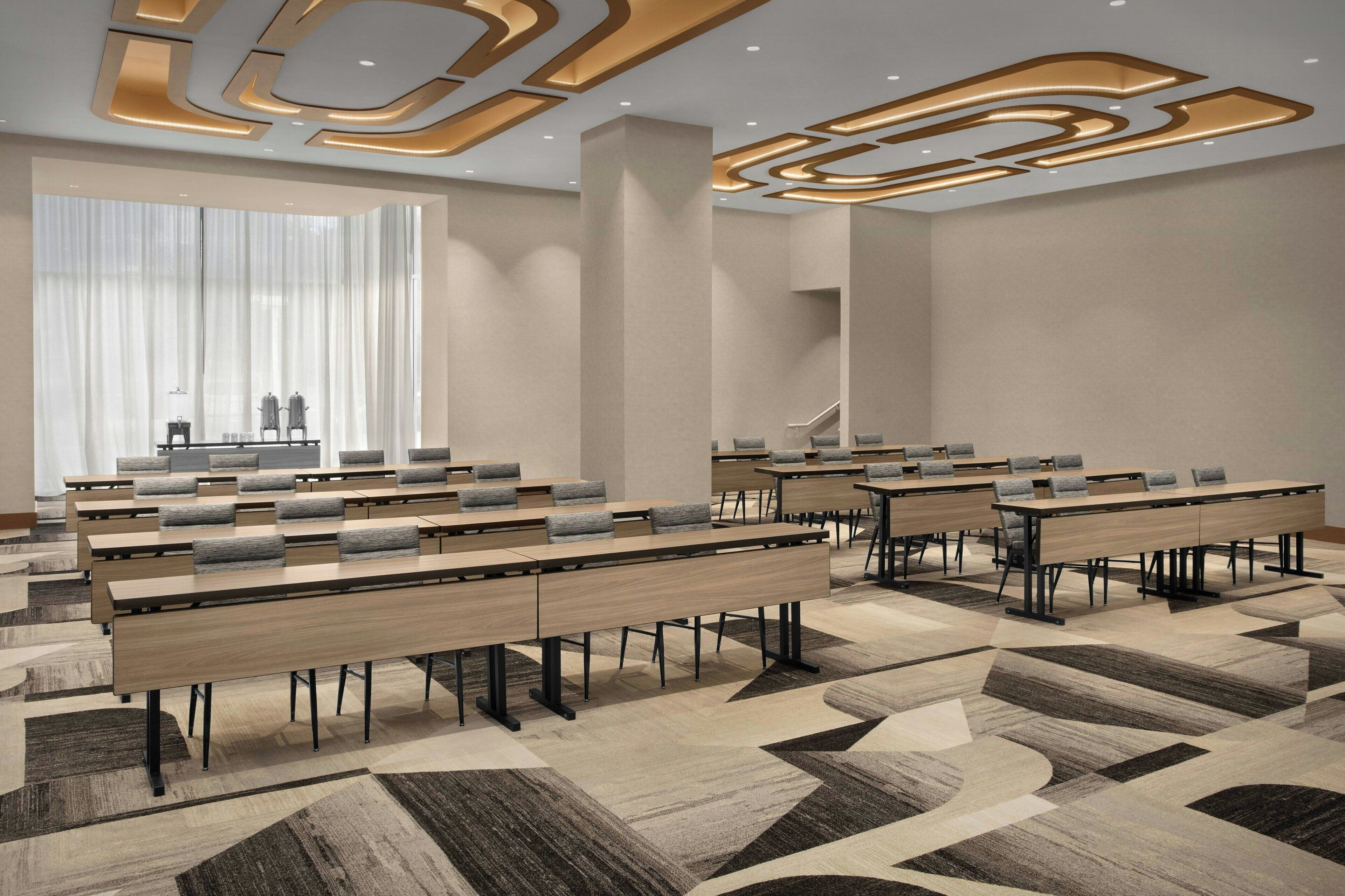 Une salle de conférence moderne, dotée de longues tables, de chaises et d’une grande fenêtre ornée de rideaux légers.