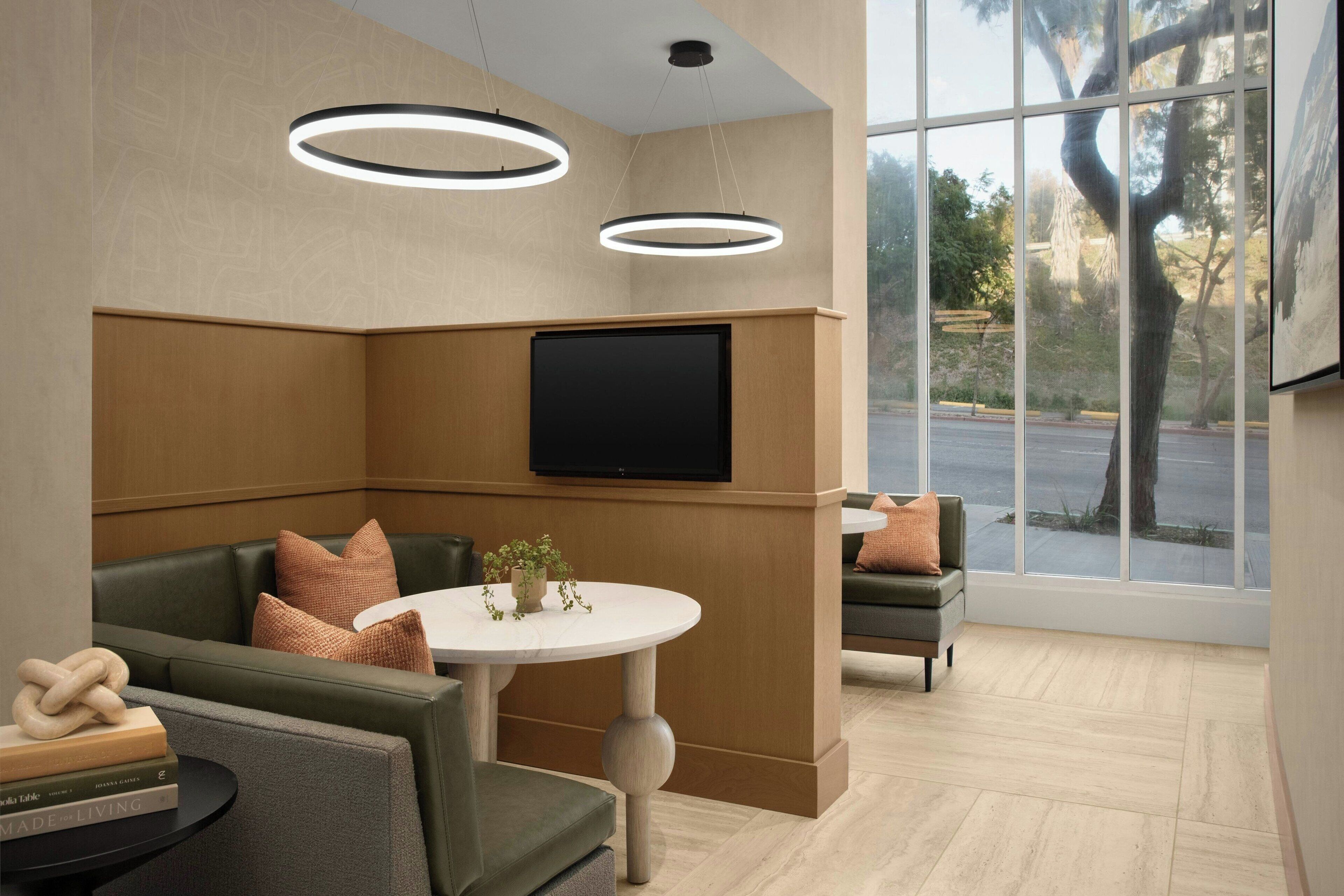 A modern waiting area with a flat-screen TV mounted on the wall, a round table with a potted plant, and a seating area with cushioned chairs and sofas.