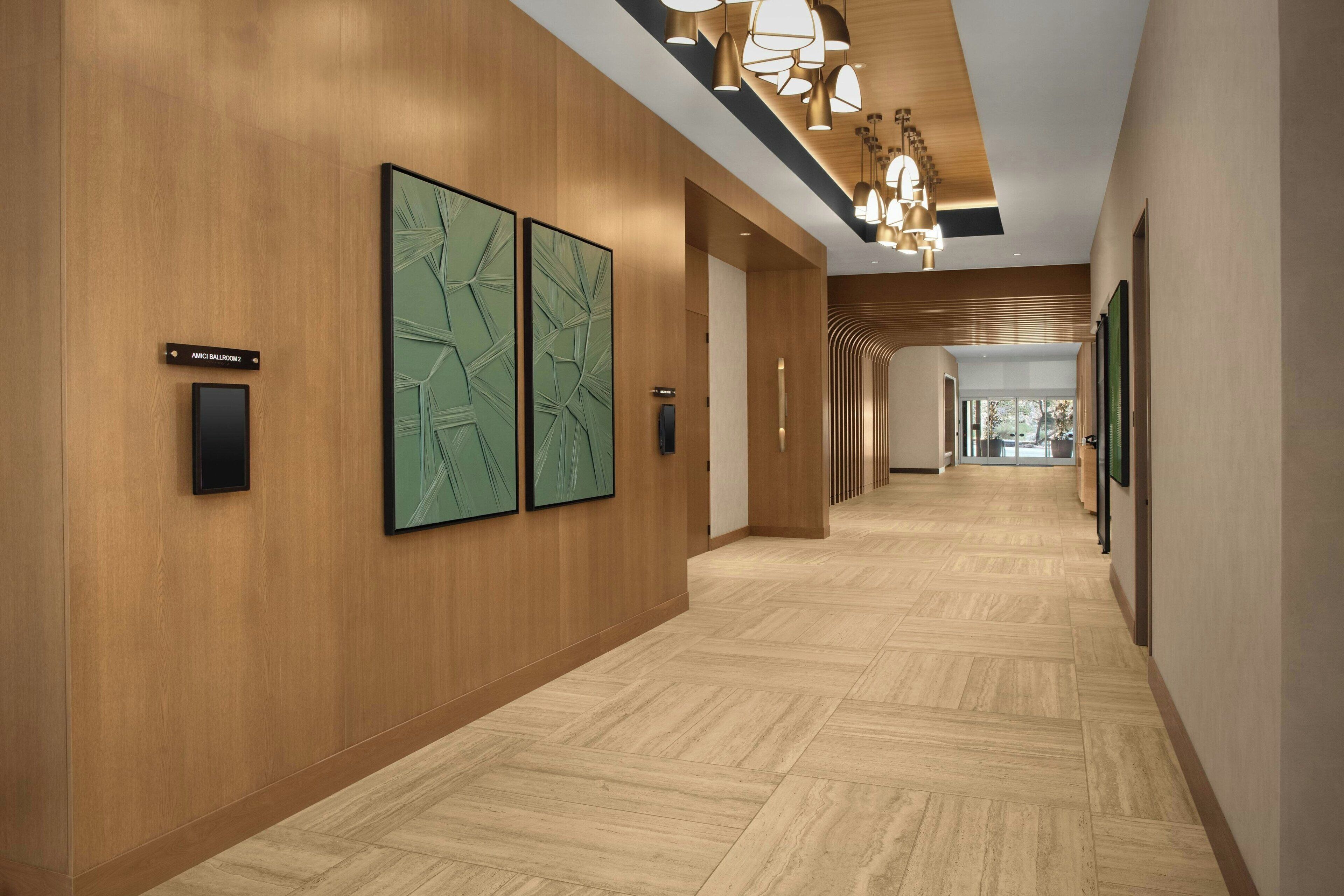 A modern hotel corridor with wooden panel walls, a wooden floor, and a ceiling with pendant lighting. There are two framed artworks on the wall and a sign indicating the location.