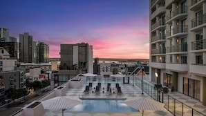 A rooftop pool area with lounge chairs and umbrellas, surrounded by modern buildings and a cityscape at sunset.