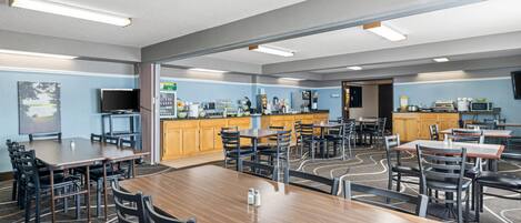 A spacious dining area with multiple tables and chairs, a kitchen area with appliances, and a television mounted on the wall.
