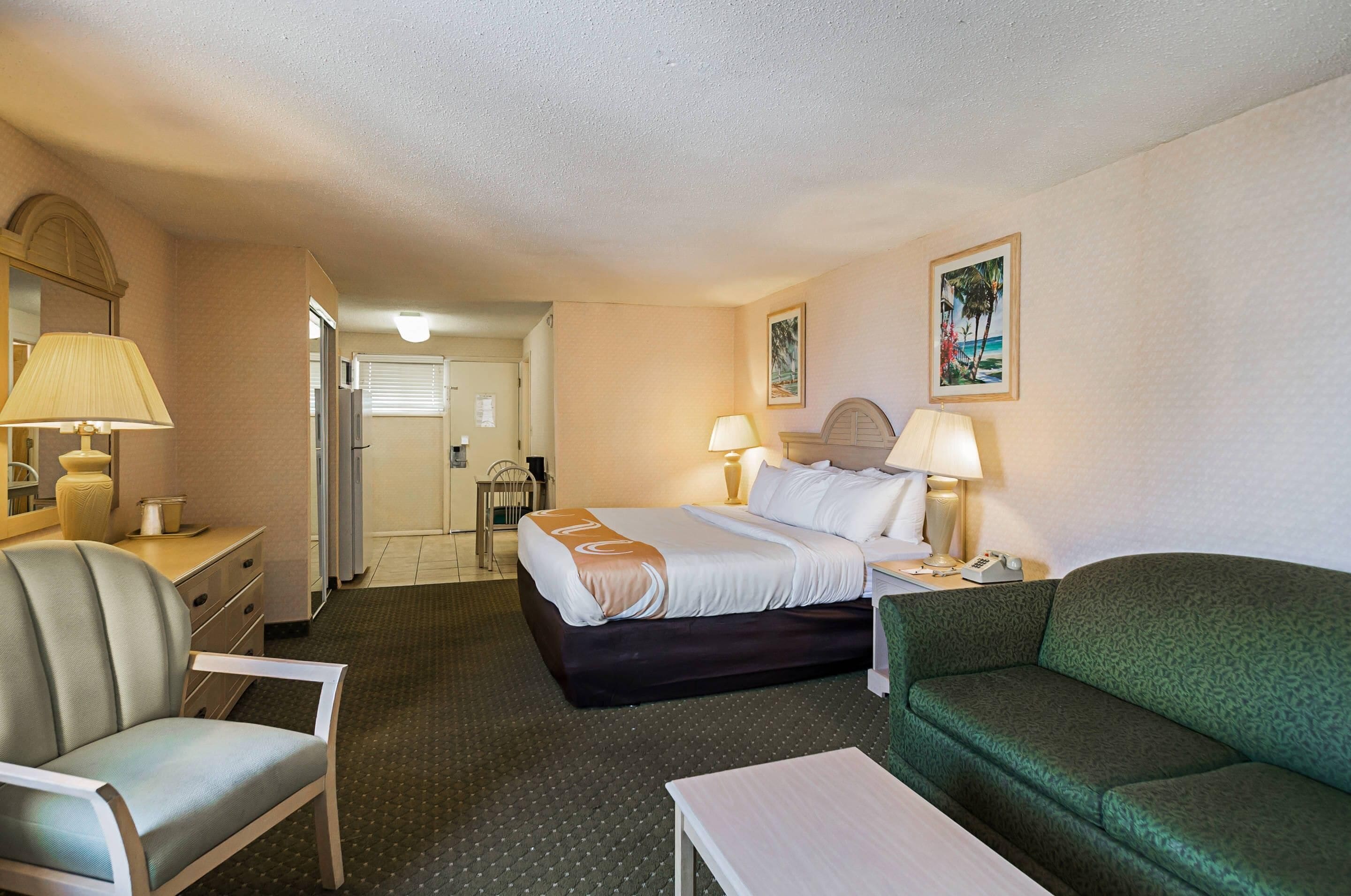 A hotel room with a bed, two bedside lamps, a green sofa, a chair, a small table, a desk with a lamp, and a mirror.
