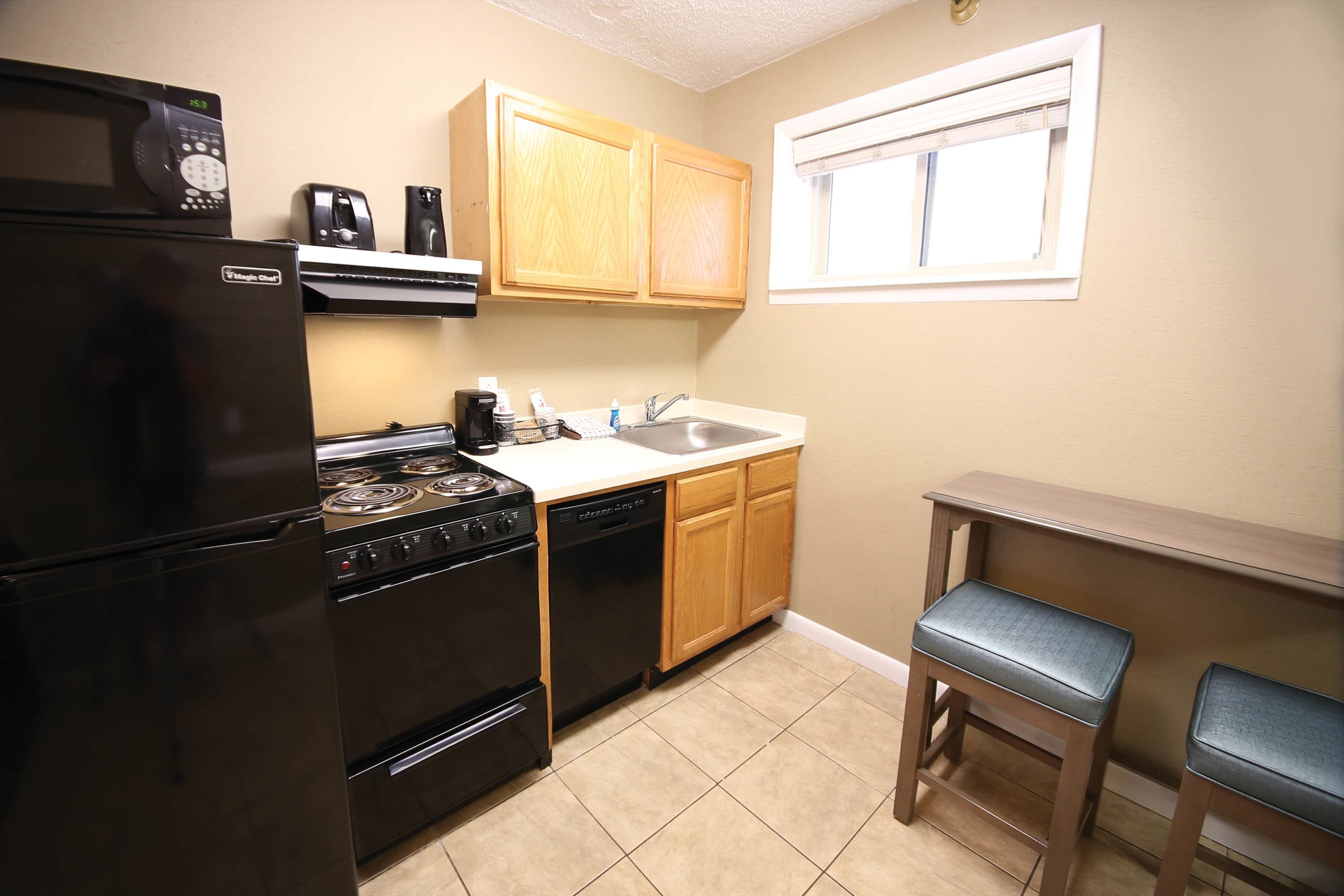 A compact kitchen with a refrigerator, stove, dishwasher, wooden cabinets, a sink, and two bar stools.