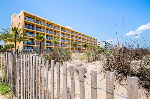 A multi-story hotel with balconies, located near a sandy beach with sparse vegetation.