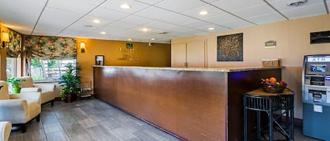 Un área de recepción con mostrador de madera, zona de asientos con sillas, frutero sobre un taburete y un cajero automático.