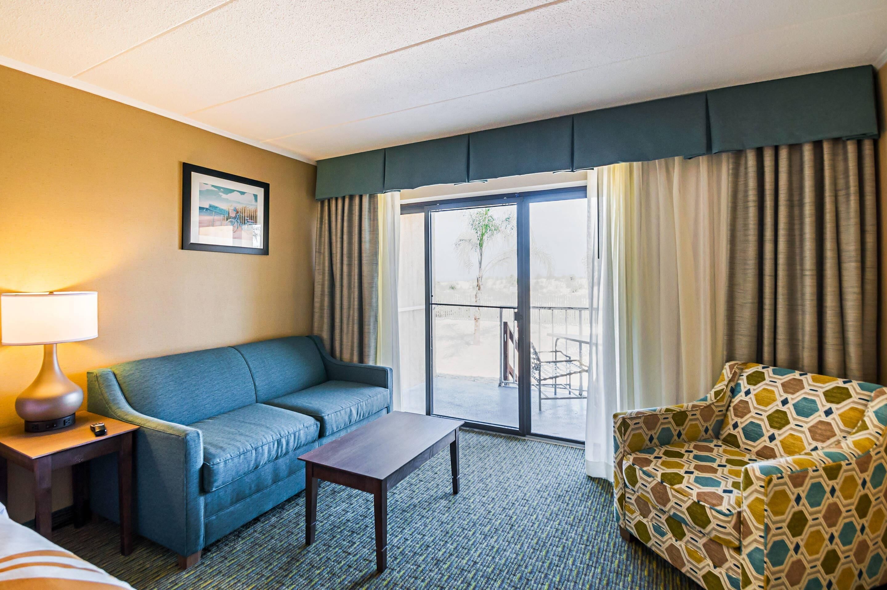 A hotel room with a blue sofa, a patterned armchair, a small table, a lamp, and a large window with curtains.