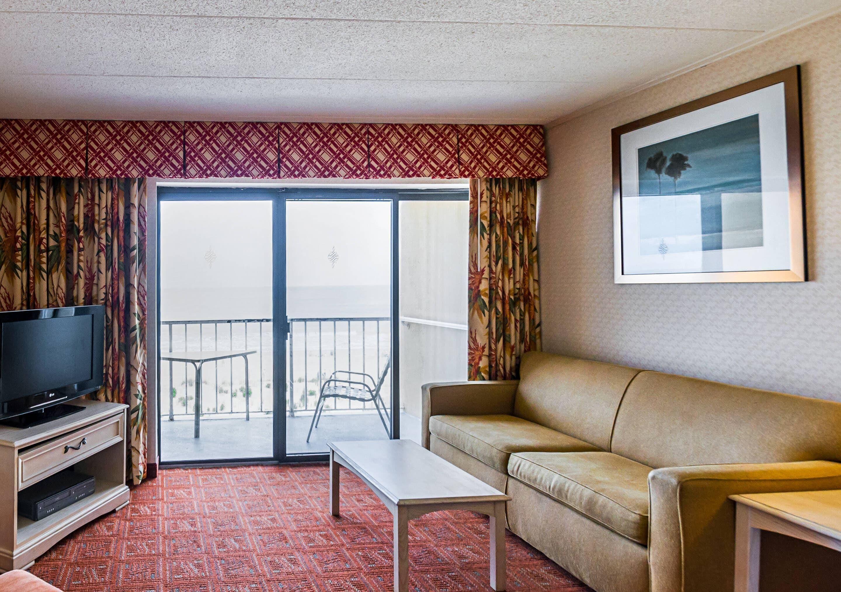 Habitación de hotel con sofá, televisor en un mueble bajo, una mesita y balcón con vistas.