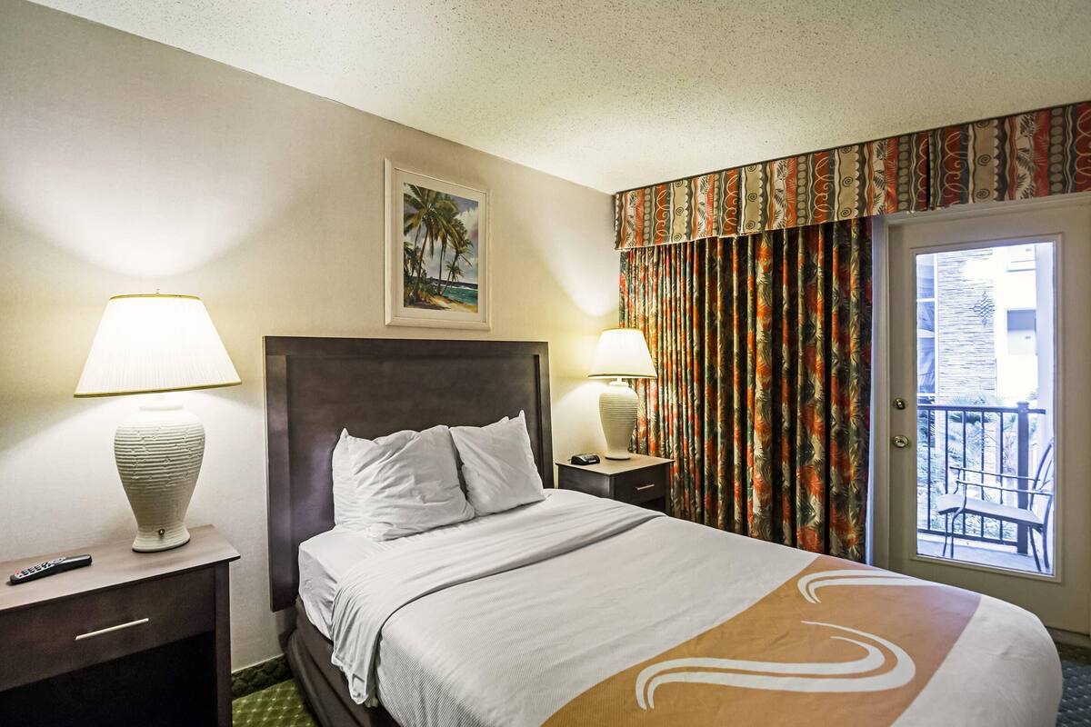 Camera d'albergo con un letto, due abat-jour, un comodino, un quadro incorniciato appeso al muro e un balcone con tende.