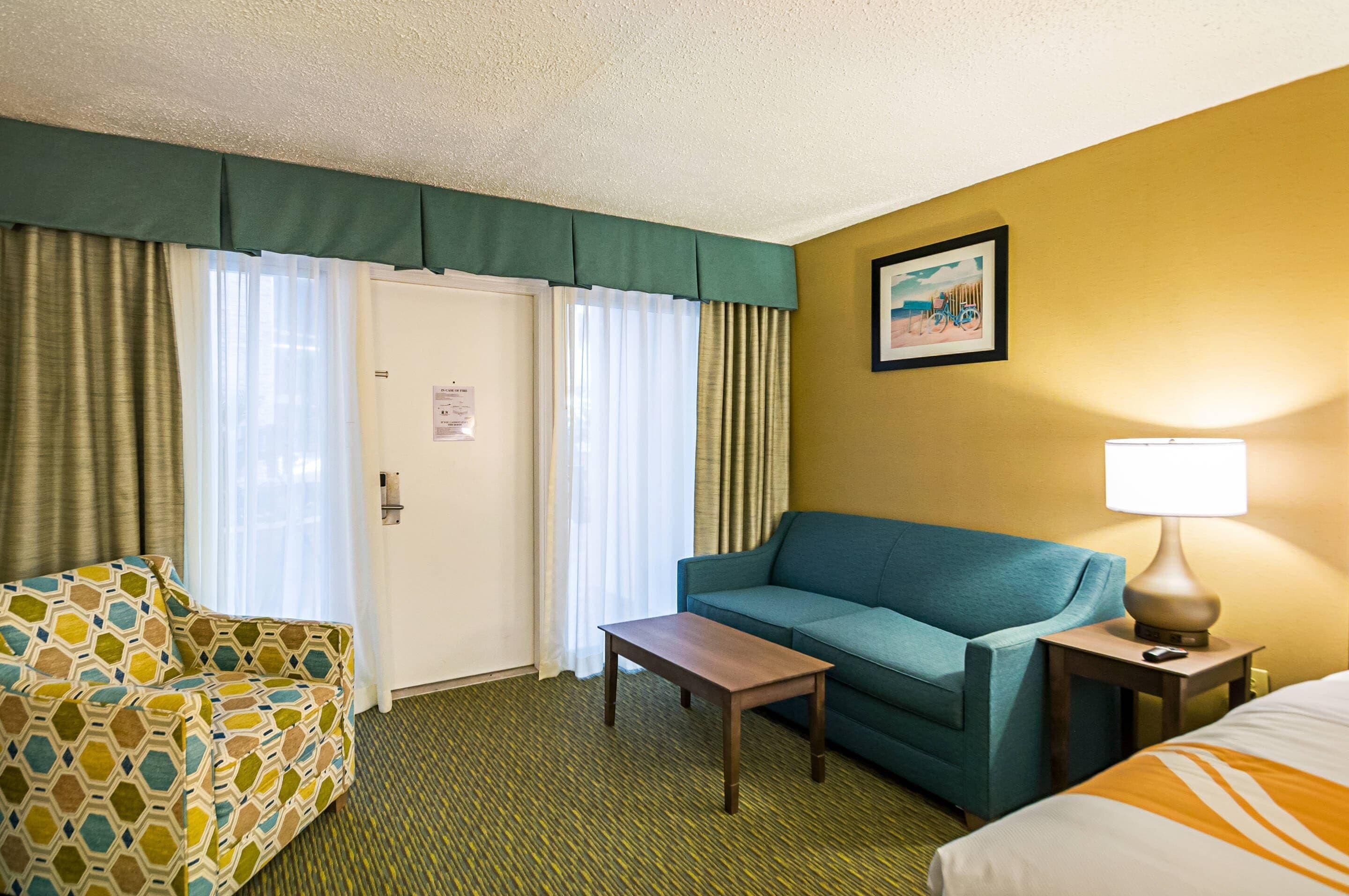 A hotel room with a green patterned sofa, a blue sofa, a wooden table, and a lamp on a bedside table.