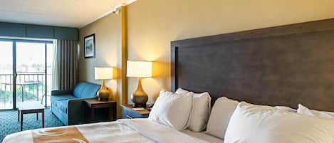 A hotel room with a large bed, a sofa, two bedside lamps, a nightstand, and a view of the outdoors through a sliding door.