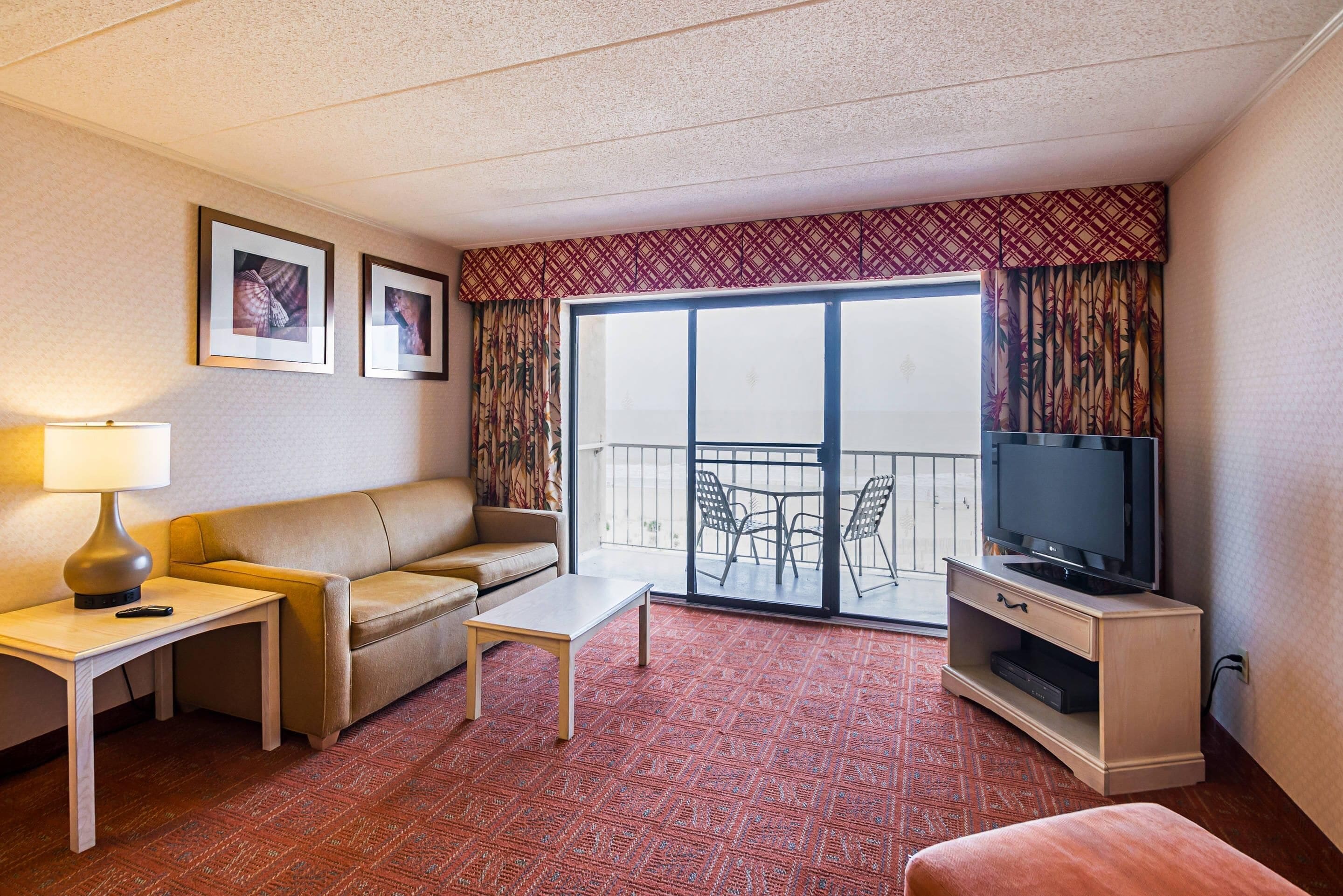 A hotel room with a sofa, a small table, a TV on a stand, and a balcony with a view.