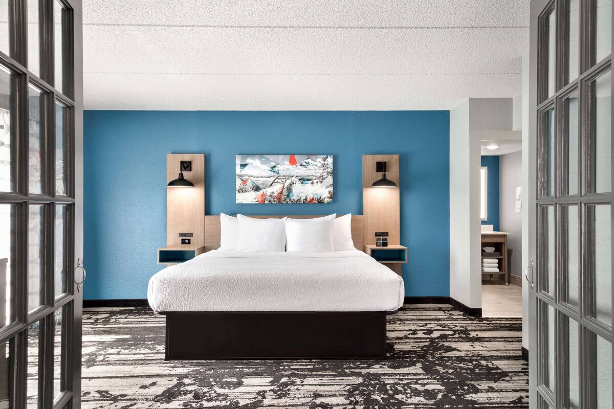 A hotel room with a large bed, blue walls, two wall-mounted lamps, and a painting above the bed.
