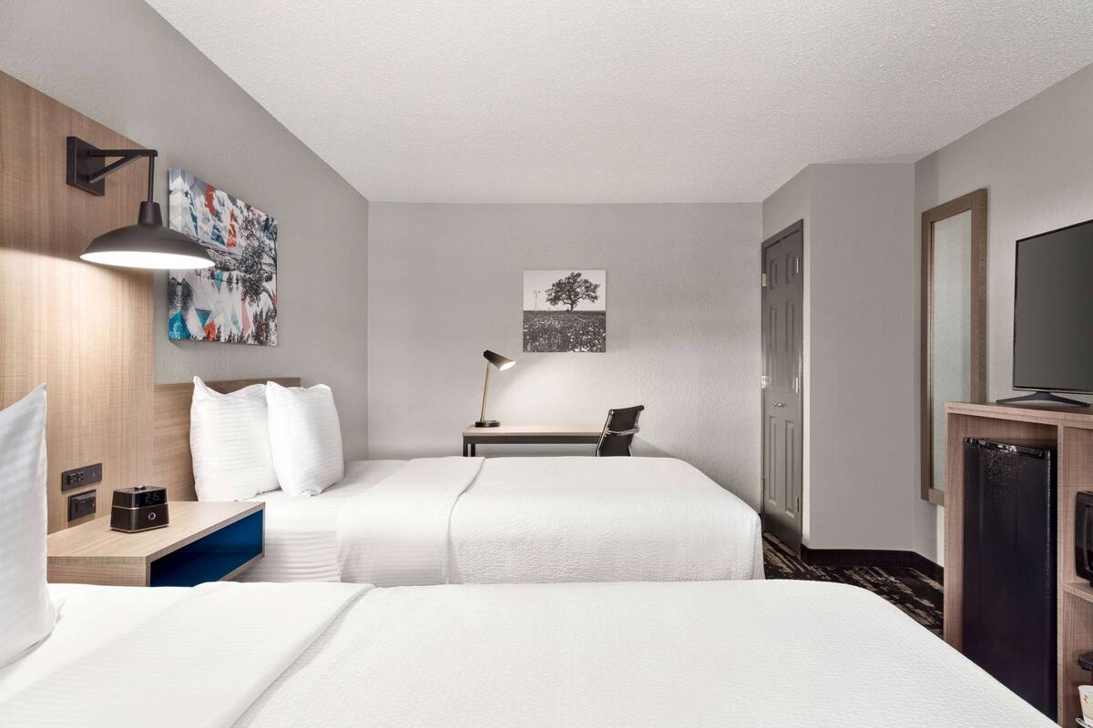 A hotel room with two beds, a desk, a chair, a TV, and artwork on the walls.