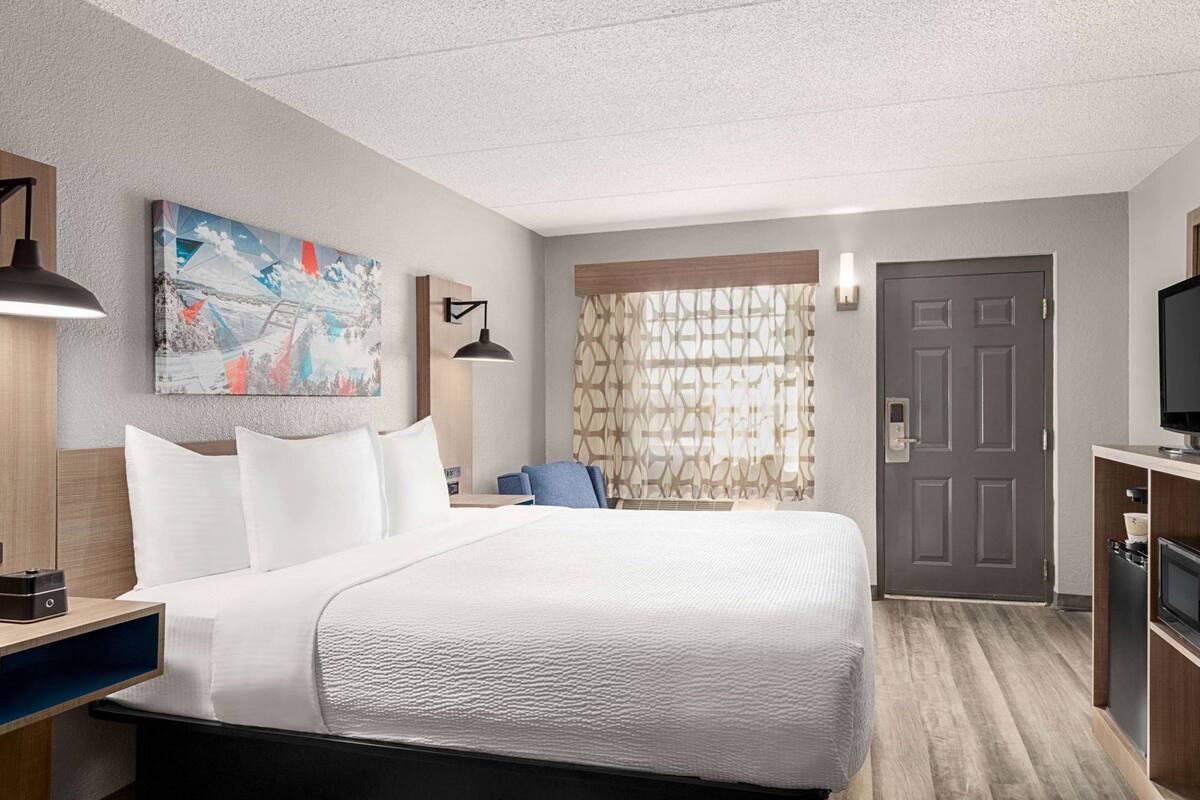 A hotel room with a bed, a desk with a lamp, a television, a painting on the wall, and a window with curtains.