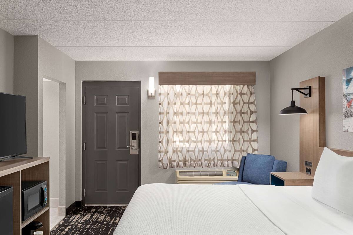 A hotel room with a bed, a TV, a desk, a chair, and a window with patterned curtains.