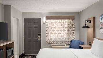 A hotel room with a bed, a TV, a desk, a chair, and a window with patterned curtains.