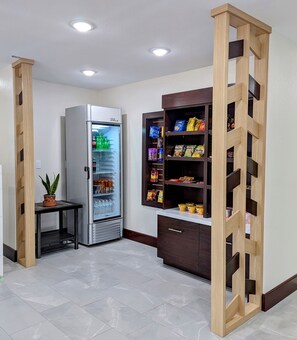 A kitchenette with a refrigerator, a microwave, a wooden cabinet with snacks, and a small table with a plant.