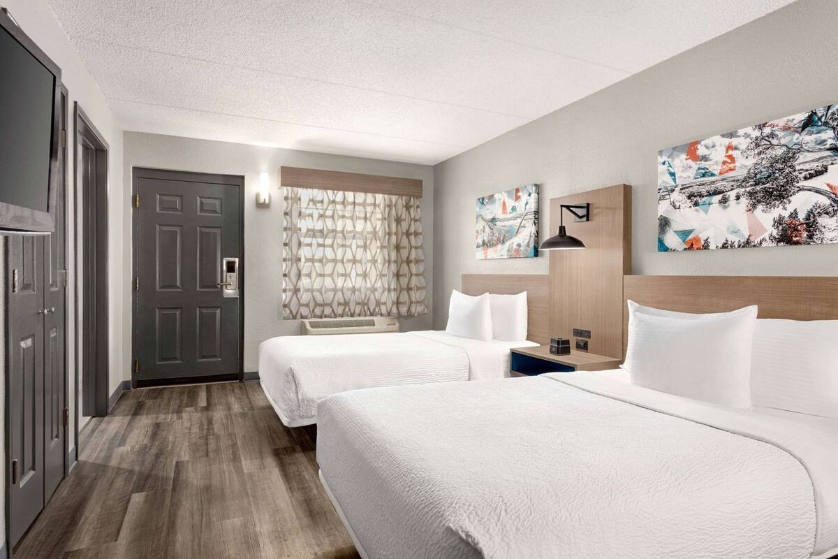 A hotel room with two beds, a wardrobe, a window with patterned curtains, and a wall-mounted artwork.