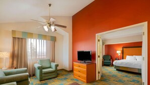 A hotel room with a bed, a television on a wooden dresser, a green armchair, a sofa, a desk, and a patterned carpet.