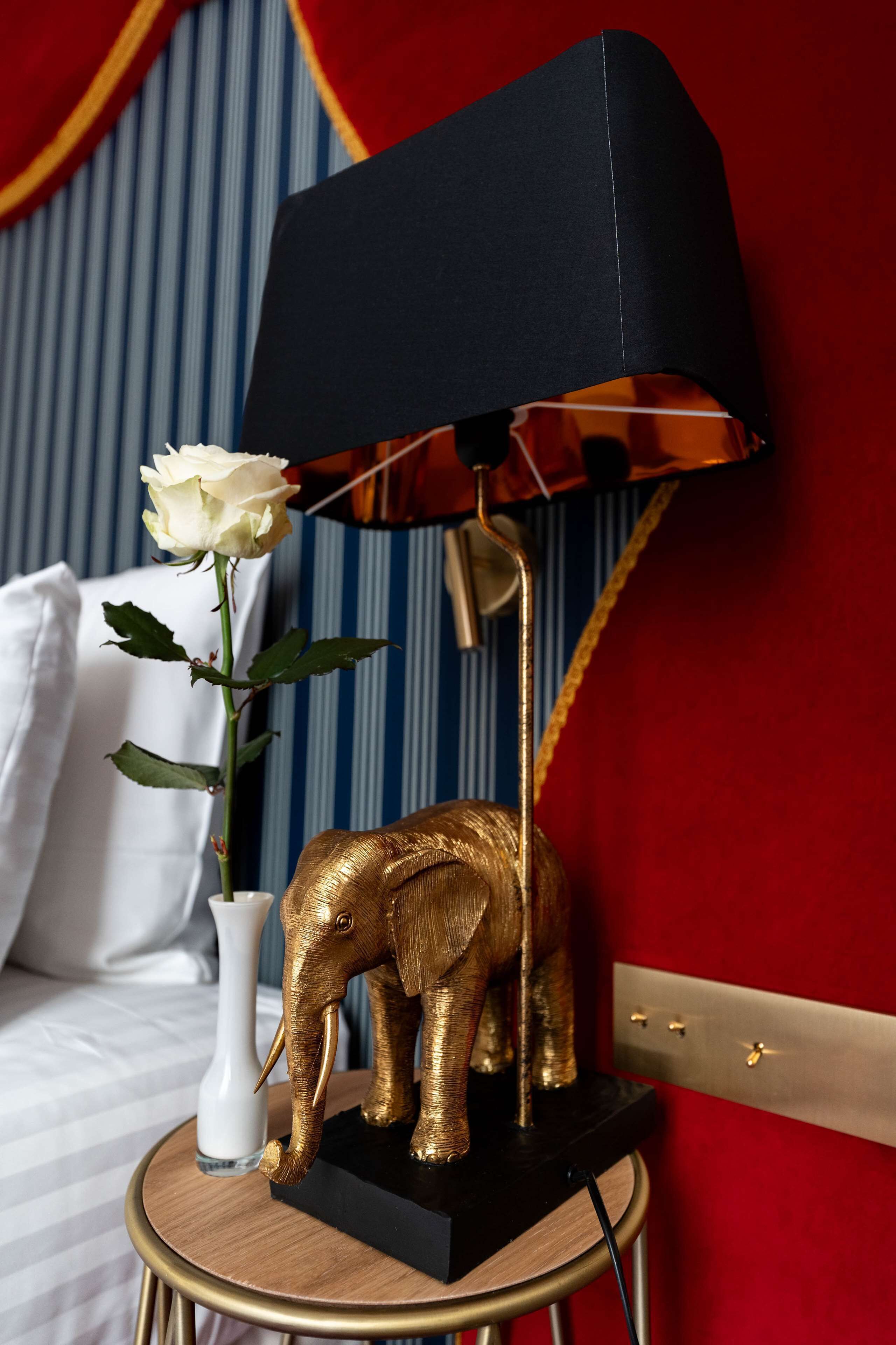 Une table de chevet ornée d’une statuette d’éléphant dorée, d’un vase blanc contenant une rose et d’une lampe noire avec un abat-jour en cuivre.