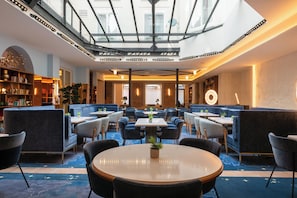En modern hotellobby med ett glastak, sittgrupper och ett stort bord i mitten med en växt.