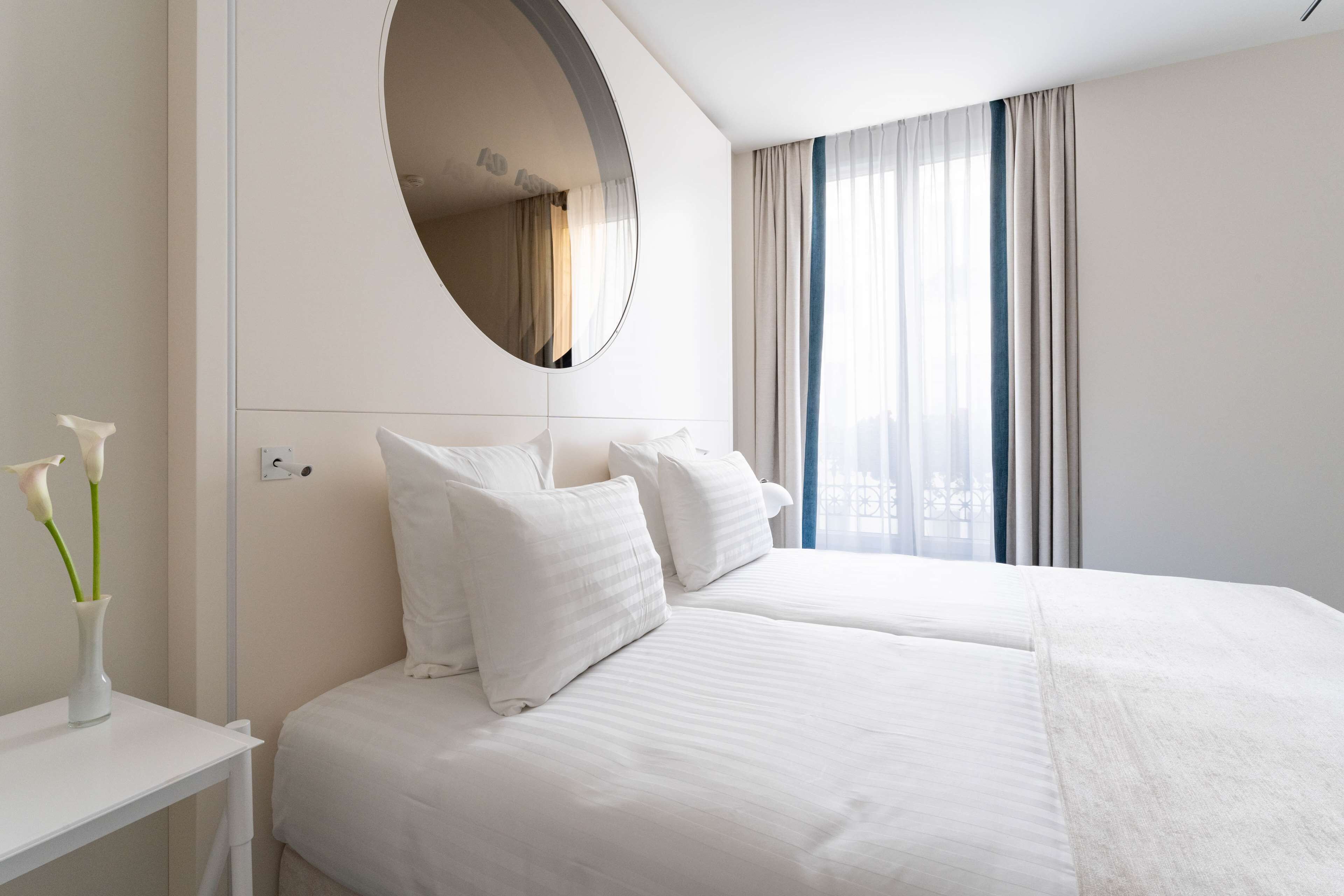 A hotel room with two beds, a round mirror, a bedside table with a vase of flowers, and a window with curtains.