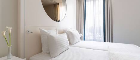 A hotel room with two beds, a round mirror, a bedside table with a vase of flowers, and a window with curtains.