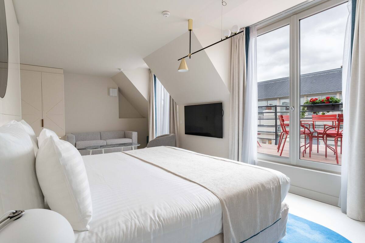 Un dormitorio moderno con una cama grande, un televisor montado en la pared y un balcón con sillas rojas.