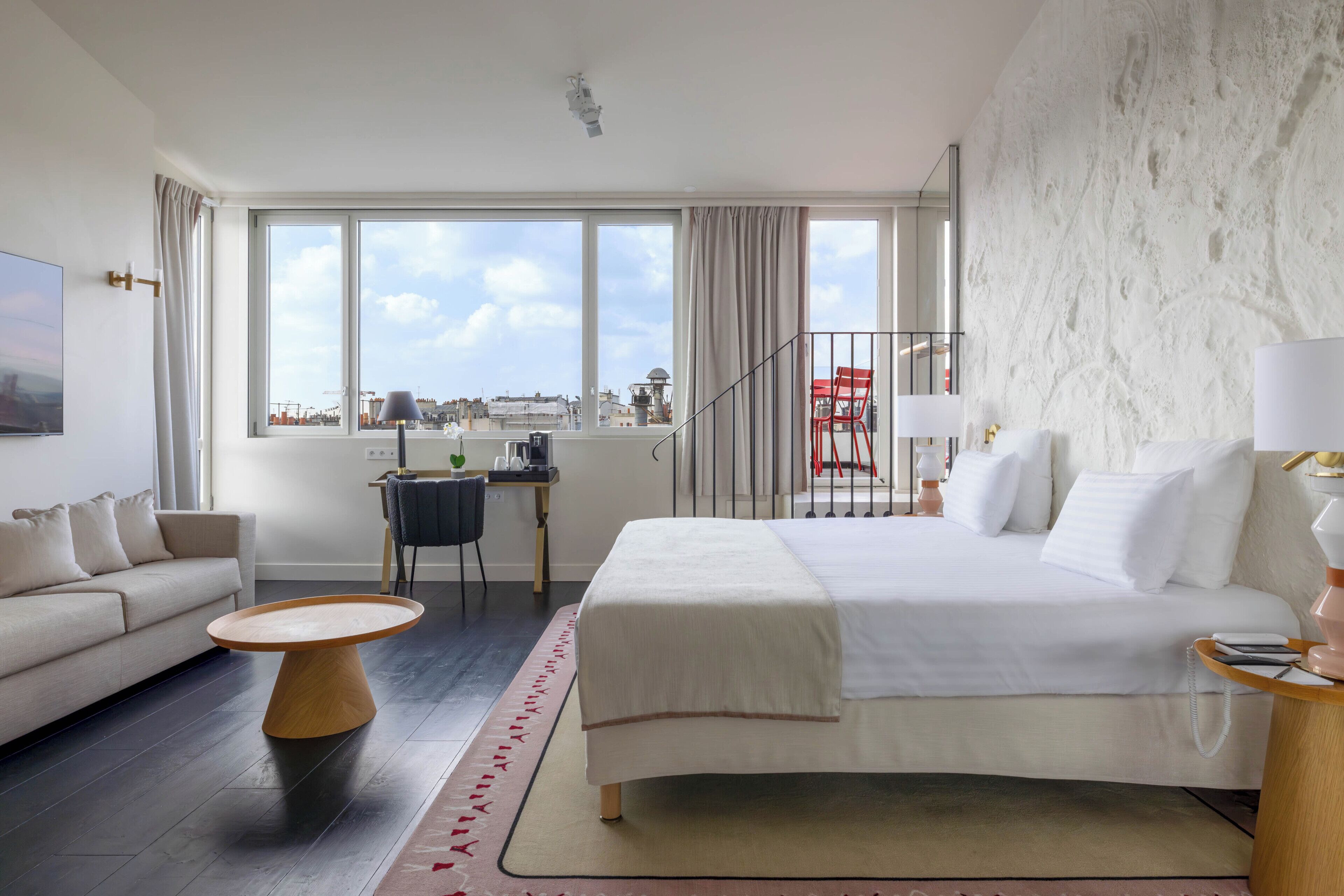 A modern hotel room with a large bed, a sofa, a dining table, and a view of the city through large windows.