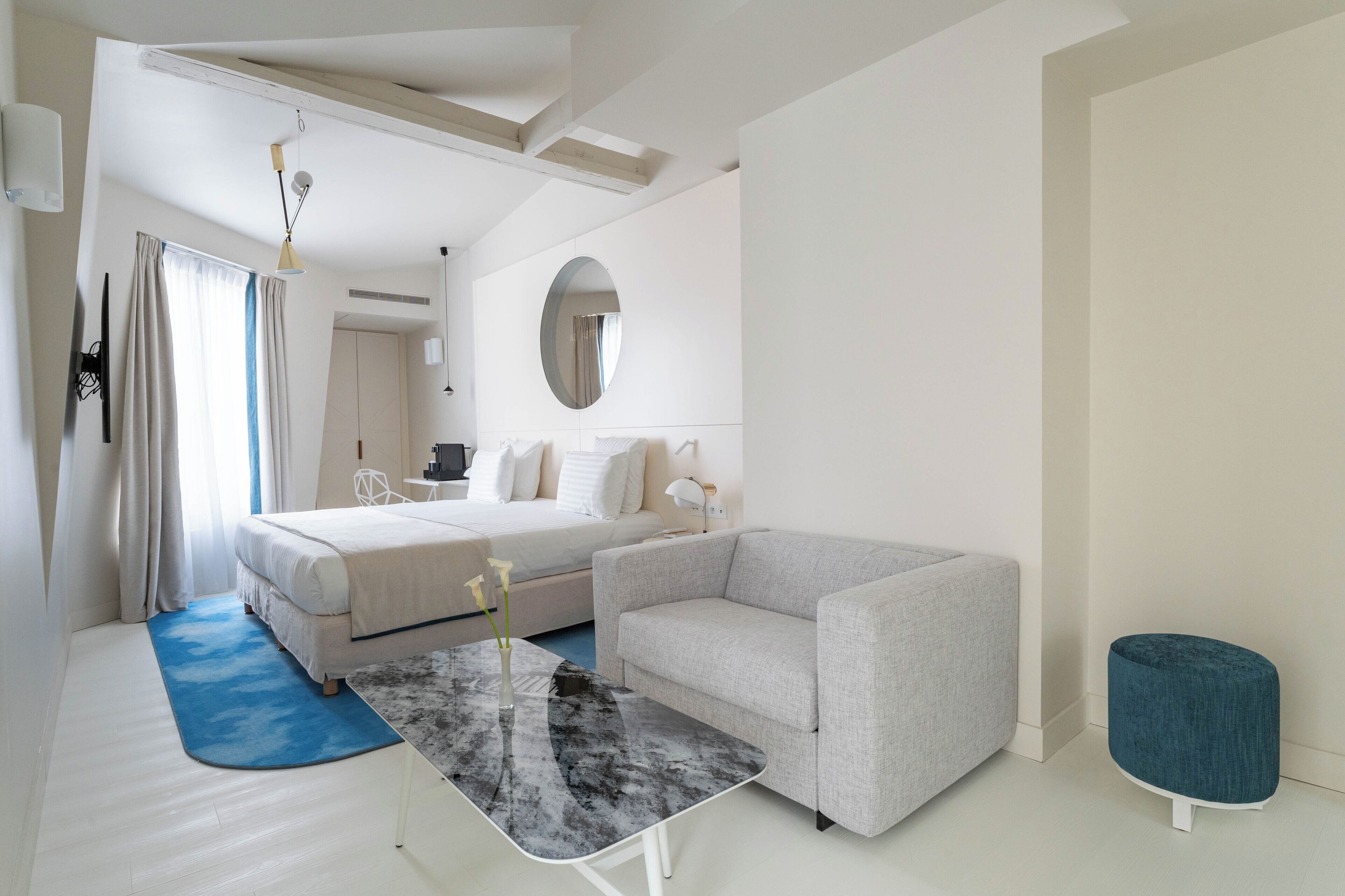 A modern hotel room with a bed, a grey sofa, a small table, a round ottoman, and a large mirror.
