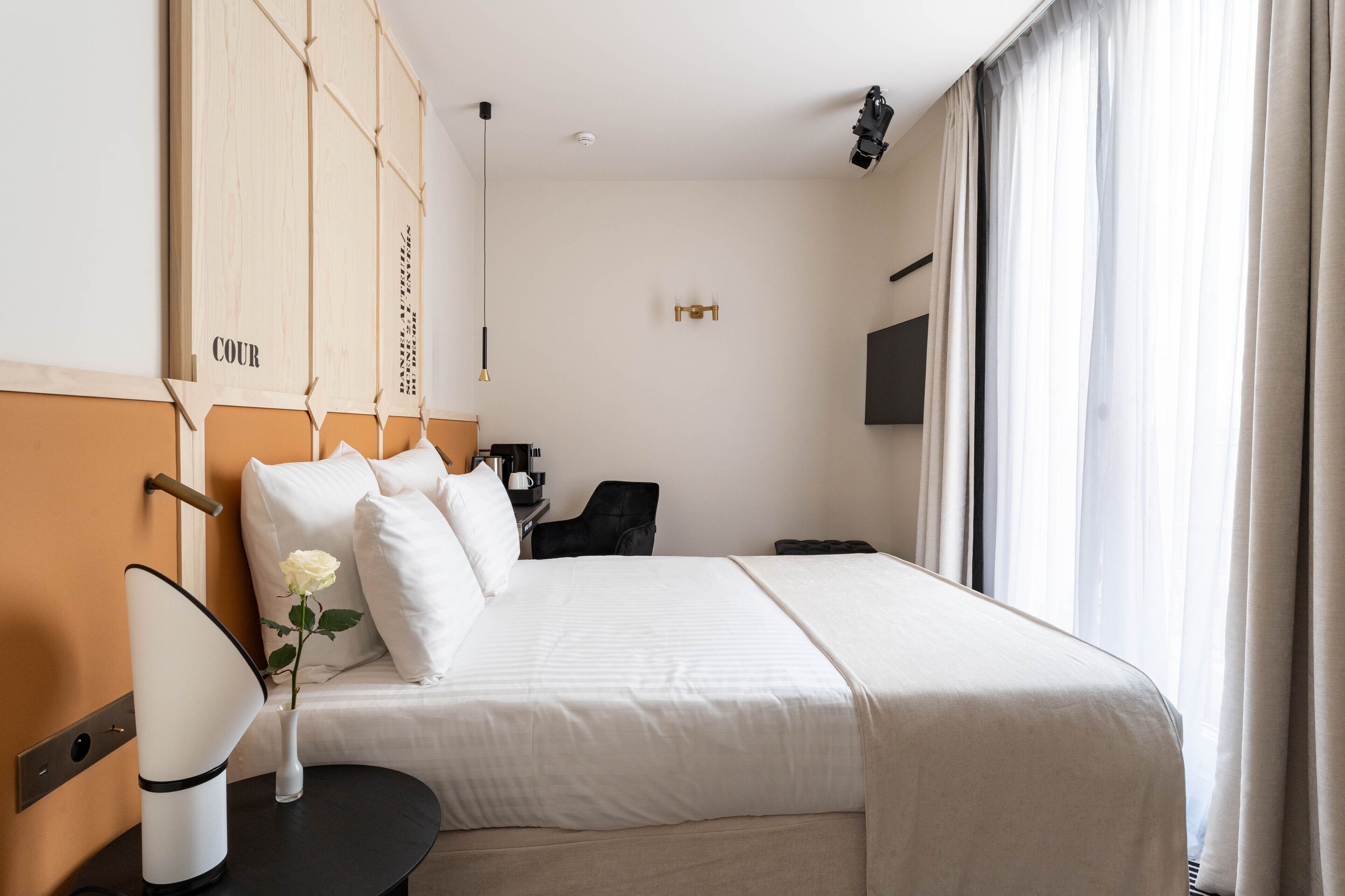 Habitación de hotel con una cama grande, una mesita de noche con lámpara, un jarrón con una flor y vistas al exterior a través de la ventana.