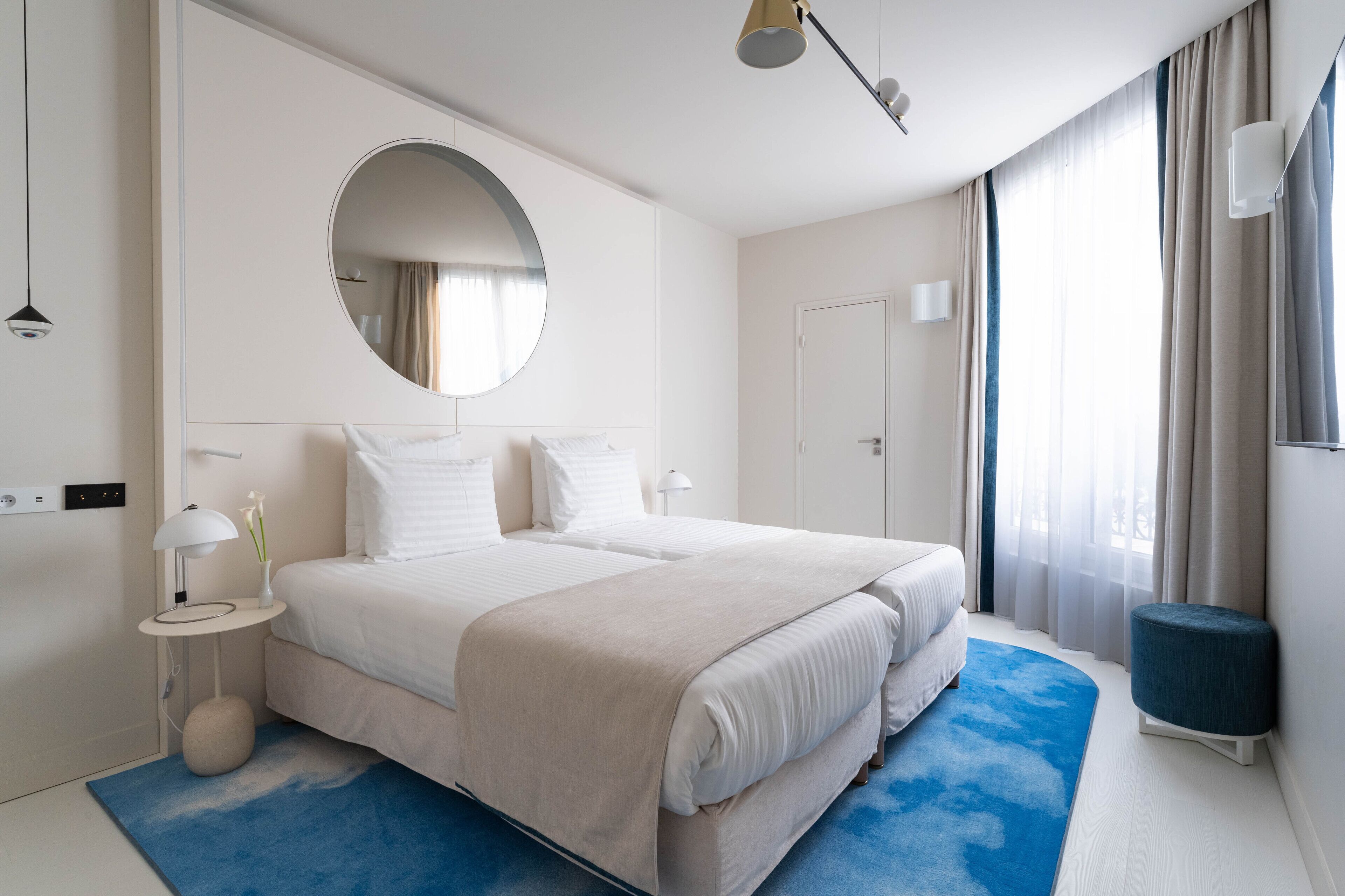 Habitación de hotel moderna con una cama grande, un espejo redondo, una mesita auxiliar pequeña y un tapete azul decorativo.