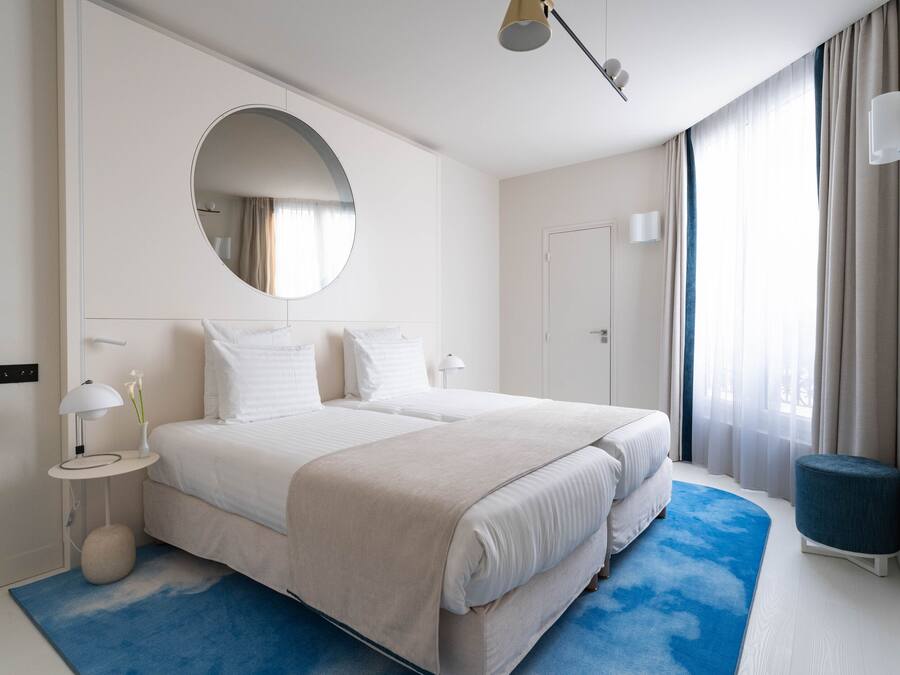 Habitación de hotel moderna con una cama grande, un espejo redondo, una mesita auxiliar pequeña y un tapete azul decorativo.