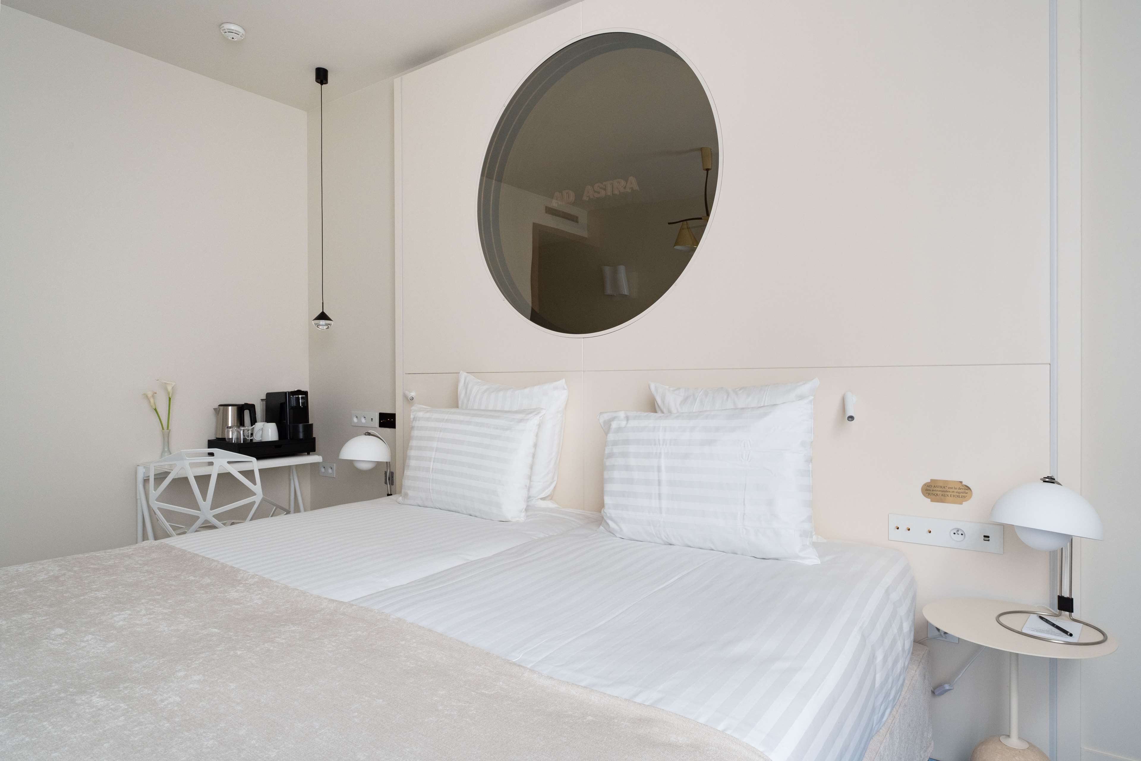 Une chambre moderne avec un grand lit, un miroir rond, une table de chevet avec une lampe et une petite table blanche sur laquelle se trouve une machine à café.